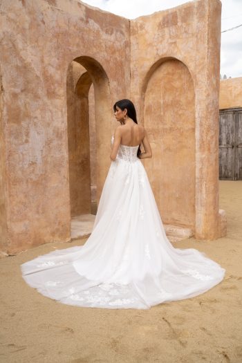 A woman wearing the GISELLE/JC2649 strapless white wedding gown with a long train stands before textured arched sandstone walls in a desert setting.