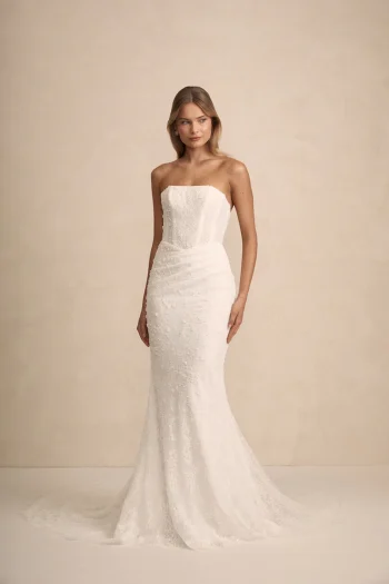 A woman stands against a plain beige background, wearing the strapless, form-fitting HAVEN by Made with Love wedding dress, featuring a subtle textured pattern and slight train.