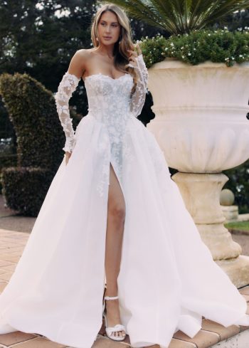 A woman in the ARTEMIS/AW2583, a white off-the-shoulder wedding gown with floral details, high slit, and sheer sleeves, stands outdoors next to a large planter.