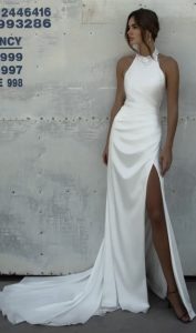 A woman models the WALSH white halter-neck gown with a high slit, standing against a metallic industrial wall marked with stenciled numbers.