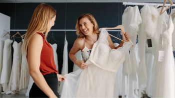 Two women stand in a bridal shop, one holding up a white wedding dress on a hanger; several other Alena Leena Bridal gowns hang in the background, highlighting this designer spotlight.