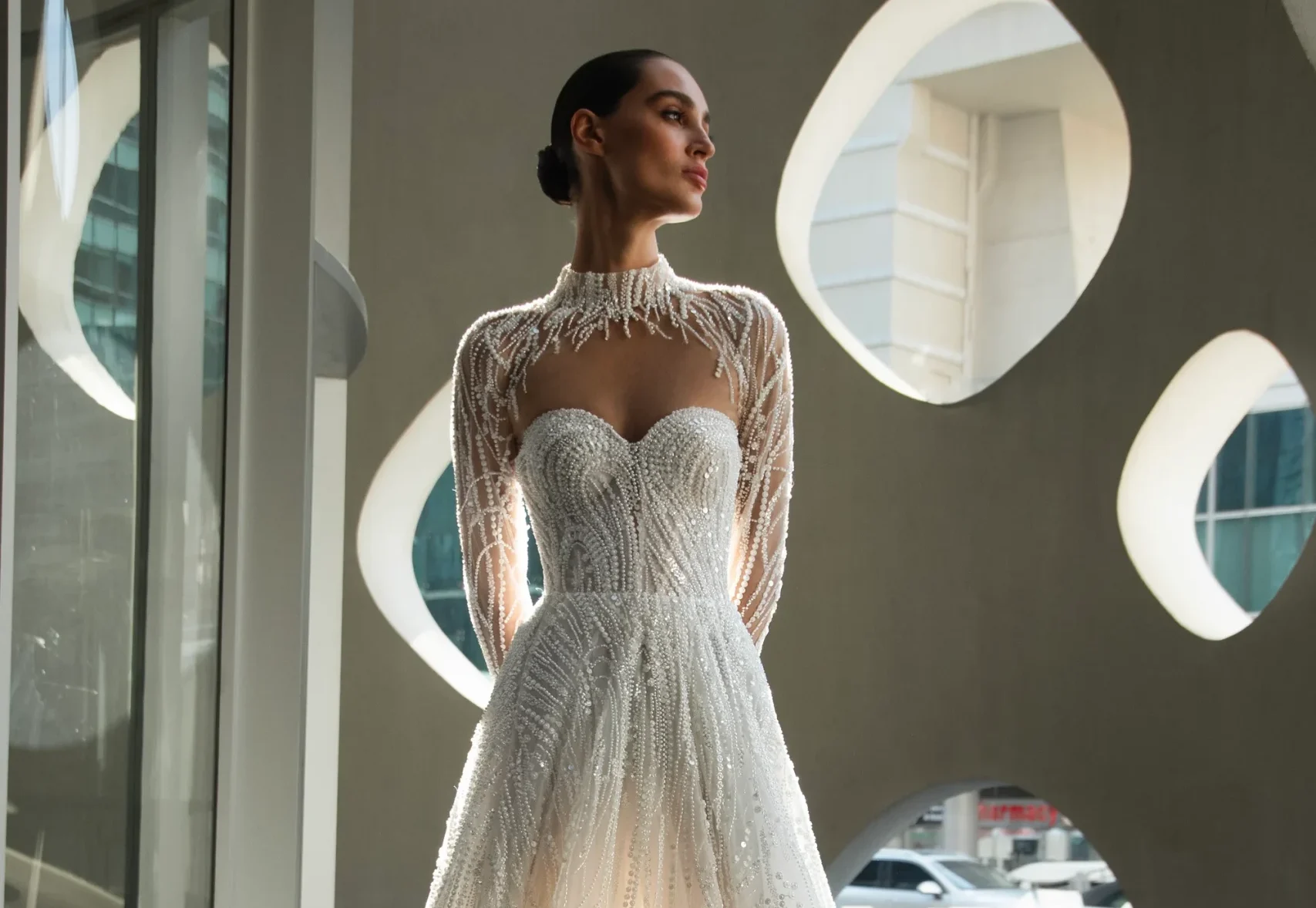 Flares Bridal + Formal | How Much Does a Wedding Dress Cost in San Francisco (2026)? A woman stands indoors by large windows, wearing the IVERA—an elegant, white, floor-length wedding gown with sheer, beaded long sleeves.