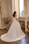 A woman in the LARISSA/88491 off-the-shoulder bridal gown by Justin Alexander stands in an ornate, vintage-style room with gold accents and decorative furniture.