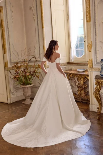 A woman wearing the LARISSA/88491 by Justin Alexander, an off-the-shoulder white bridal gown, stands indoors facing a window with her long train gracefully spread behind her.