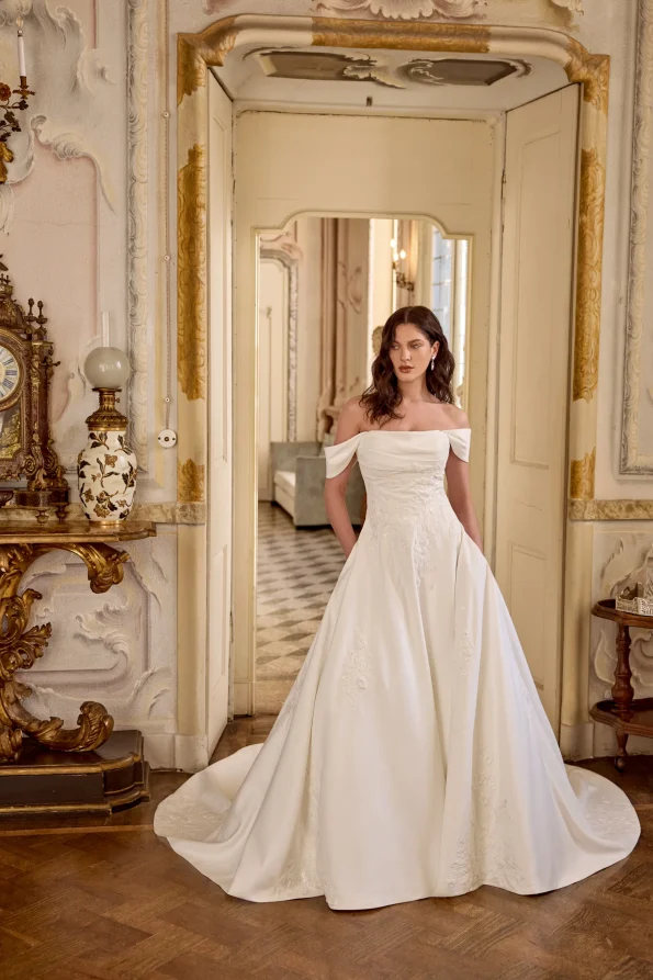 A woman in the LARISSA/88491 off-the-shoulder bridal gown by Justin Alexander stands in an ornate, vintage-style room with gold accents and decorative furniture.
