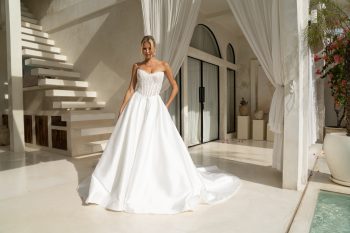 A woman stands indoors near white curtains and modern architecture, bathed in sunlight, wearing the elegant strapless KIARA/MC2701 wedding gown.