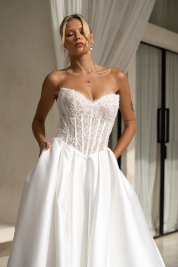 A woman poses before sheer curtains in the KIARA/MC2701 strapless white wedding dress, showcasing a lace bodice, satin skirt, and handy pockets for a modern bridal style.