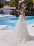 A woman in the ETERNITY by Enzoani sleeveless lace and tulle wedding dress stands poolside in the sun, framed by palm trees and white buildings, embodying timeless elegance.