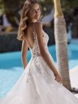 A woman in the ETERNITY by Enzoani sleeveless lace and tulle wedding dress stands poolside in the sun, framed by palm trees and white buildings, embodying timeless elegance.