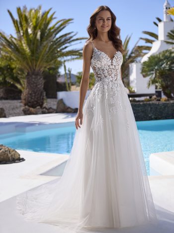 A woman in the ETERNITY by Enzoani sleeveless lace and tulle wedding dress stands poolside in the sun, framed by palm trees and white buildings, embodying timeless elegance.