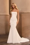 A woman stands against a neutral stone background in the M2576 by Mikaella, a strapless, fitted white wedding dress featuring a subtle train.