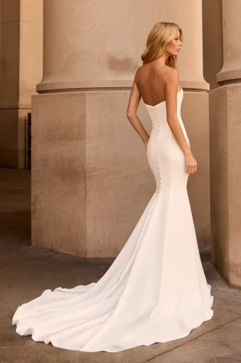 A woman stands between two large stone columns wearing the strapless, fitted M2576 by Mikaella wedding dress, featuring a long train and button details down the back.