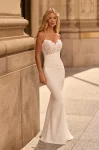 A woman stands outdoors before an ornate stone building, wearing the M2581 by Mikaella—a white sleeveless wedding dress featuring a lace bodice and a fitted satin skirt.