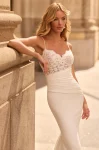 A woman stands outdoors before an ornate stone building, wearing the M2581 by Mikaella—a white sleeveless wedding dress featuring a lace bodice and a fitted satin skirt.