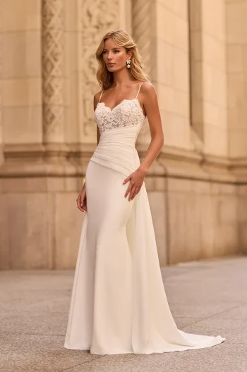 A woman stands outdoors before an ornate stone building, wearing the M2581 by Mikaella—a white sleeveless wedding dress featuring a lace bodice and a fitted satin skirt.