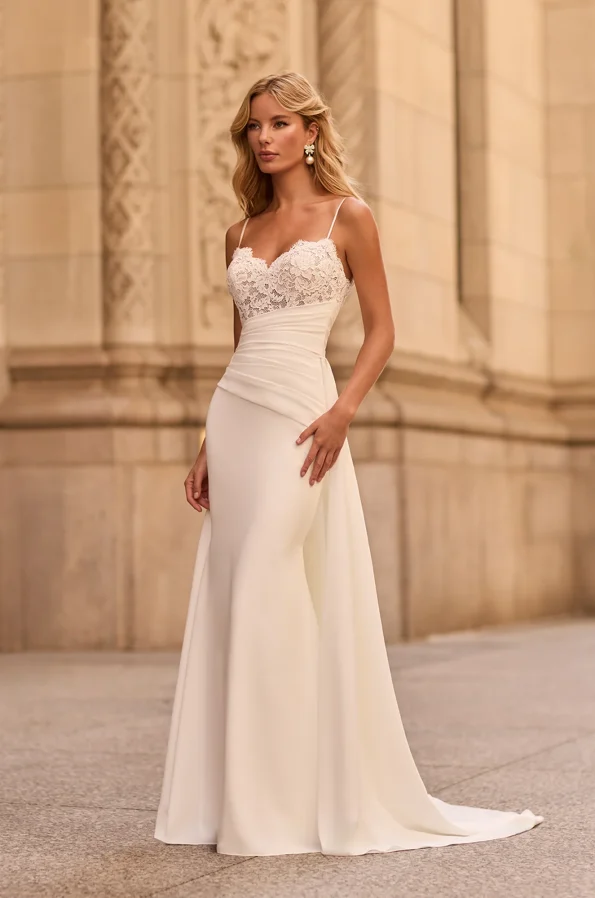 A woman stands outdoors before an ornate stone building, wearing the M2581 by Mikaella—a white sleeveless wedding dress featuring a lace bodice and a fitted satin skirt.