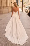 A woman stands outdoors before an ornate stone building, wearing the M2581 by Mikaella—a white sleeveless wedding dress featuring a lace bodice and a fitted satin skirt.