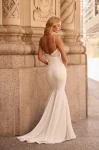 A woman stands outdoors before an ornate stone building, wearing the M2581 by Mikaella—a white sleeveless wedding dress featuring a lace bodice and a fitted satin skirt.