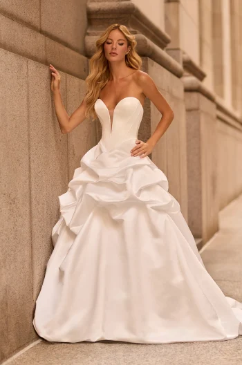 A woman in the M2584 by Mikaella, a strapless white ballgown with a deep plunging neckline and dramatic ruffled skirt, stands against a stone building wall.