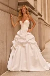 A woman in the M2584 by Mikaella, a strapless white ballgown with a deep plunging neckline and dramatic ruffled skirt, stands against a stone building wall.