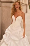 A woman in the M2584 by Mikaella, a strapless white ballgown with a deep plunging neckline and dramatic ruffled skirt, stands against a stone building wall.
