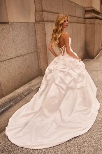 A woman in the M2584 by Mikaella, a strapless white gown with a corset back and draped skirt, stands on a stone walkway beside a grand building.
