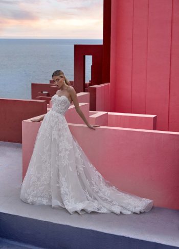 A woman wears the GINA/MJ1063 by Allure, a strapless lace wedding gown, standing against a pink architectural backdrop with the sea and sky in the distance.