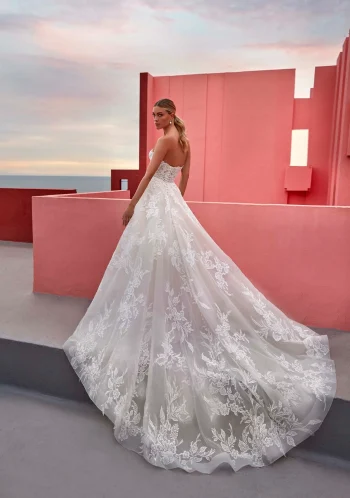 A woman in the strapless white lace GINA/MJ1063 by Allure wedding gown stands on a pink-walled rooftop at sunset, looking over her shoulder.
