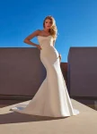 A woman radiates charm in the ERIN/ MJ1153 by Allure, a fitted strapless white gown, as she stands outdoors against a textured wall, sunlight casting graceful shadows around her.