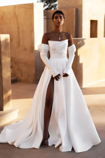 Outdoors, a woman wears the MANAMA off-the-shoulder white gown with long sleeves and a high slit, accessorized with a pendant necklace.