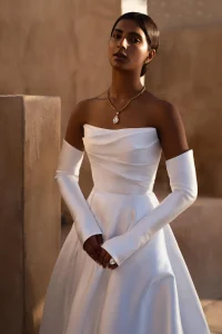 A woman in the white off-shoulder satin MANAMA gown with long gloves wears a gold necklace and ring, standing indoors against a neutral-toned background.