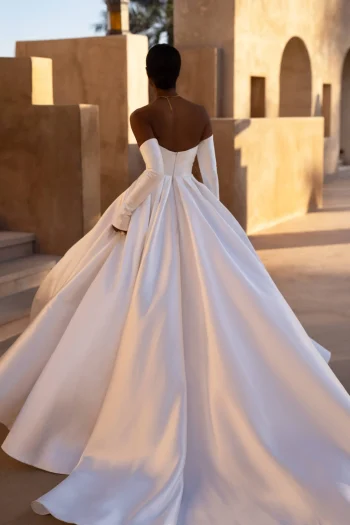 A person stands outdoors near tan stone walls wearing the MANAMA off-the-shoulder, long-sleeved white gown with a full skirt.