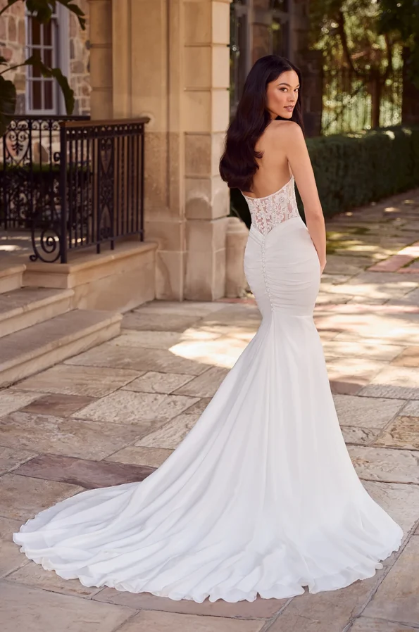 Woman in a strapless, form-fitting P5179 by Mikaella wedding dress with a long train and sheer lace back, standing outdoors on a stone patio near a building with columns and a wrought iron railing.