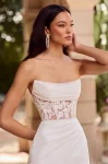 A woman stands outdoors on a stone walkway near an arch and greenery, wearing the P5179 by Mikaella—an off-the-shoulder white gown featuring a sheer lace bodice.