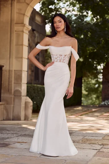 A woman stands outdoors on a stone walkway near an arch and greenery, wearing the P5179 by Mikaella—an off-the-shoulder white gown featuring a sheer lace bodice.