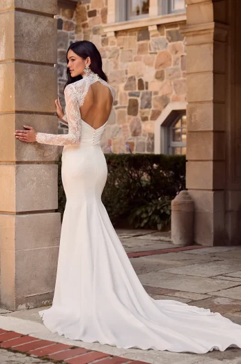 A woman in the P5185 by Mikaella bridal gown, featuring lace sleeves and an open back, stands near a stone building with her back to the camera.