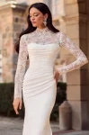 Flares Bridal + Formal | P5185 by Mikaella A woman stands outdoors before a stone building, wearing the Mikaella P5185—a long, fitted white gown featuring lace sleeves and a high lace neckline.