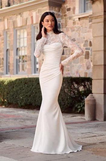 A woman stands outdoors before a stone building, wearing the Mikaella P5185—a long, fitted white gown featuring lace sleeves and a high lace neckline.