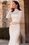 Flares Bridal + Formal | P5185 by Mikaella A woman stands outdoors before a stone building, wearing the Mikaella P5185—a long, fitted white gown featuring lace sleeves and a high lace neckline.