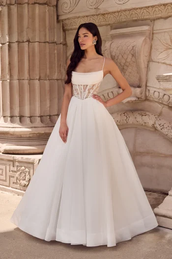 A woman stands outdoors in front of a stone wall, wearing the P5186 by Mikaella—a white sleeveless wedding dress with a fitted, semi-sheer corset bodice and full floor-length skirt.
