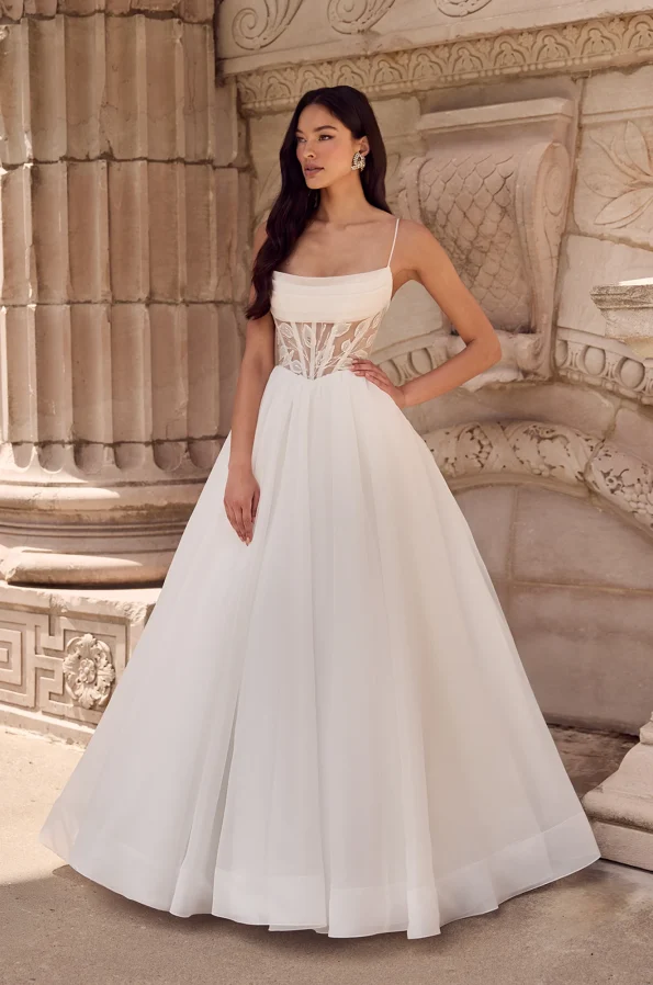 A woman stands outdoors in front of a stone wall, wearing the P5186 by Mikaella—a white sleeveless wedding dress with a fitted, semi-sheer corset bodice and full floor-length skirt.