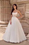 A woman stands outdoors in front of a stone wall, wearing the P5186 by Mikaella—a white sleeveless wedding dress with a fitted, semi-sheer corset bodice and full floor-length skirt.