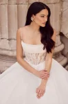 A woman stands outdoors in front of a stone wall, wearing the P5186 by Mikaella—a white sleeveless wedding dress with a fitted, semi-sheer corset bodice and full floor-length skirt.