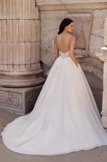 A woman faces away from the camera in front of ornate stone architecture, wearing the backless P5186 by Mikaella wedding gown with a long train.