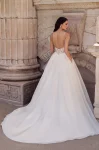 A woman stands outdoors in front of a stone wall, wearing the P5186 by Mikaella—a white sleeveless wedding dress with a fitted, semi-sheer corset bodice and full floor-length skirt.