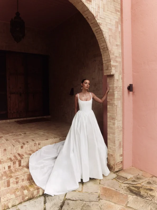 A woman in a white, floor-length Alena Lenna gown with a long train stands under a brick archway by a pink wall, holding the PROTEA by Alena Lenna bouquet.