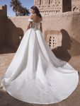A woman stands outdoors wearing TEMINA by Enzoani, a stunning white off-the-shoulder bridal gown featuring a fitted bodice and long train.