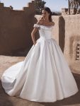 A woman stands outdoors wearing TEMINA by Enzoani, a stunning white off-the-shoulder bridal gown featuring a fitted bodice and long train.