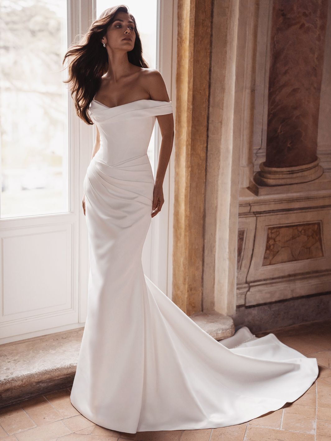 A woman in the off-the-shoulder, fitted TRISHA by Enzoani wedding dress with a train stands indoors near a large window and ornate stone walls. A woman in the off-the-shoulder, fitted TRISHA by Enzoani wedding dress with a train stands indoors near a large window and ornate stone walls.