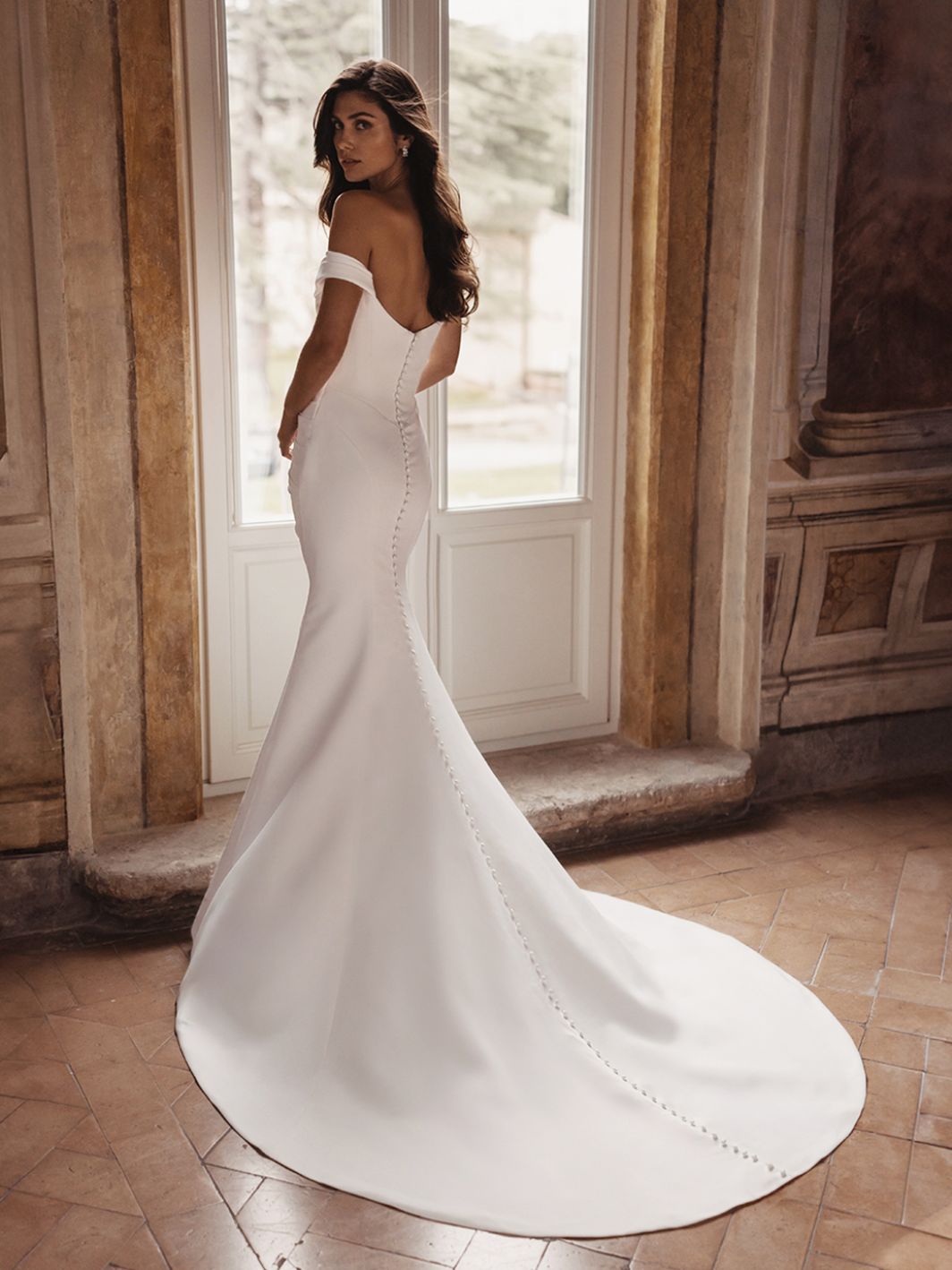 A woman wearing the off-the-shoulder, fitted TRISHA by Enzoani bridal gown with a long train and button accents stands beside a large window in an ornate room. A woman wearing the off-the-shoulder, fitted TRISHA by Enzoani bridal gown with a long train and button accents stands beside a large window in an ornate room.